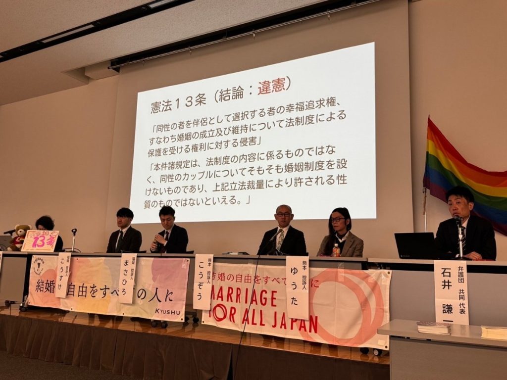 報告会の様子。原告さん4名と弁護団共同代表が壇上に並んでいる。投影スライドには、次のことが書かれている。
憲法13条　結論違憲
「同性の者を伴侶として選択する者の幸福追求権、すなわち婚姻の成立及び維持について法制度による保護を受ける権利に対する侵害」
「本件諸規定は、法制度の内容に係るものではなく、同性のカップルについてそもそも婚姻制度を設けないものであり、上記立法裁量により許される性質のものではないといえる。」
